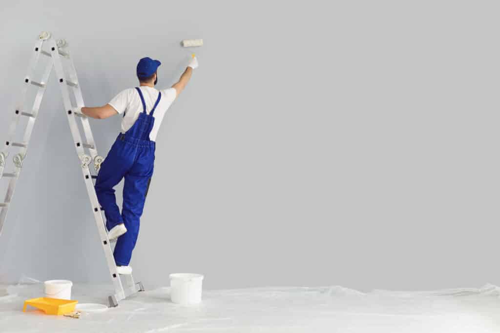 Peintre en combinaison bleue sur une échelle, dos tourné, appliquant de la peinture claire sur un mur avec un rouleau. Seau et plateau au sol protégé.