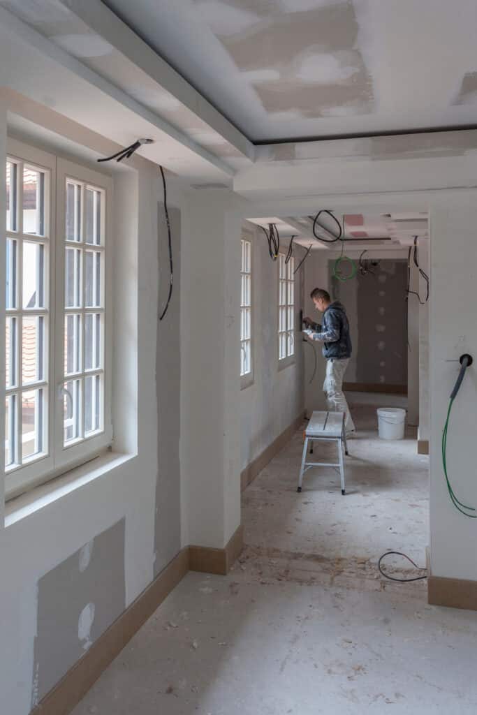 Ouvrier enduit un mur près de fenêtres dans une pièce en rénovation. Câbles électriques au plafond, sol de chantier poussiéreux.